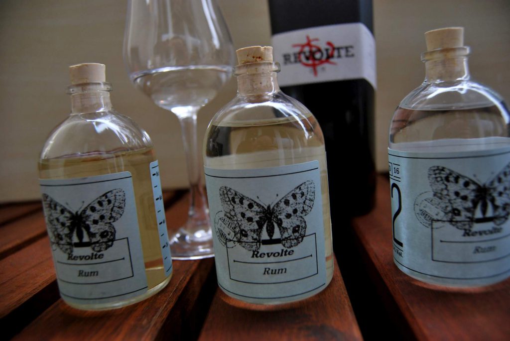 Dreimal Revolte Rum, dreimal unterschiedliche Farben. Im Hintergrund der Original-Rum aus der Original-Flasche.