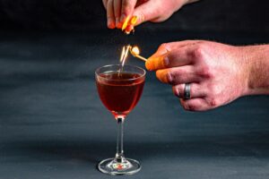 Ein Mancini Cocktail, der mit einer Orangenzeste abgeflämmt wird.